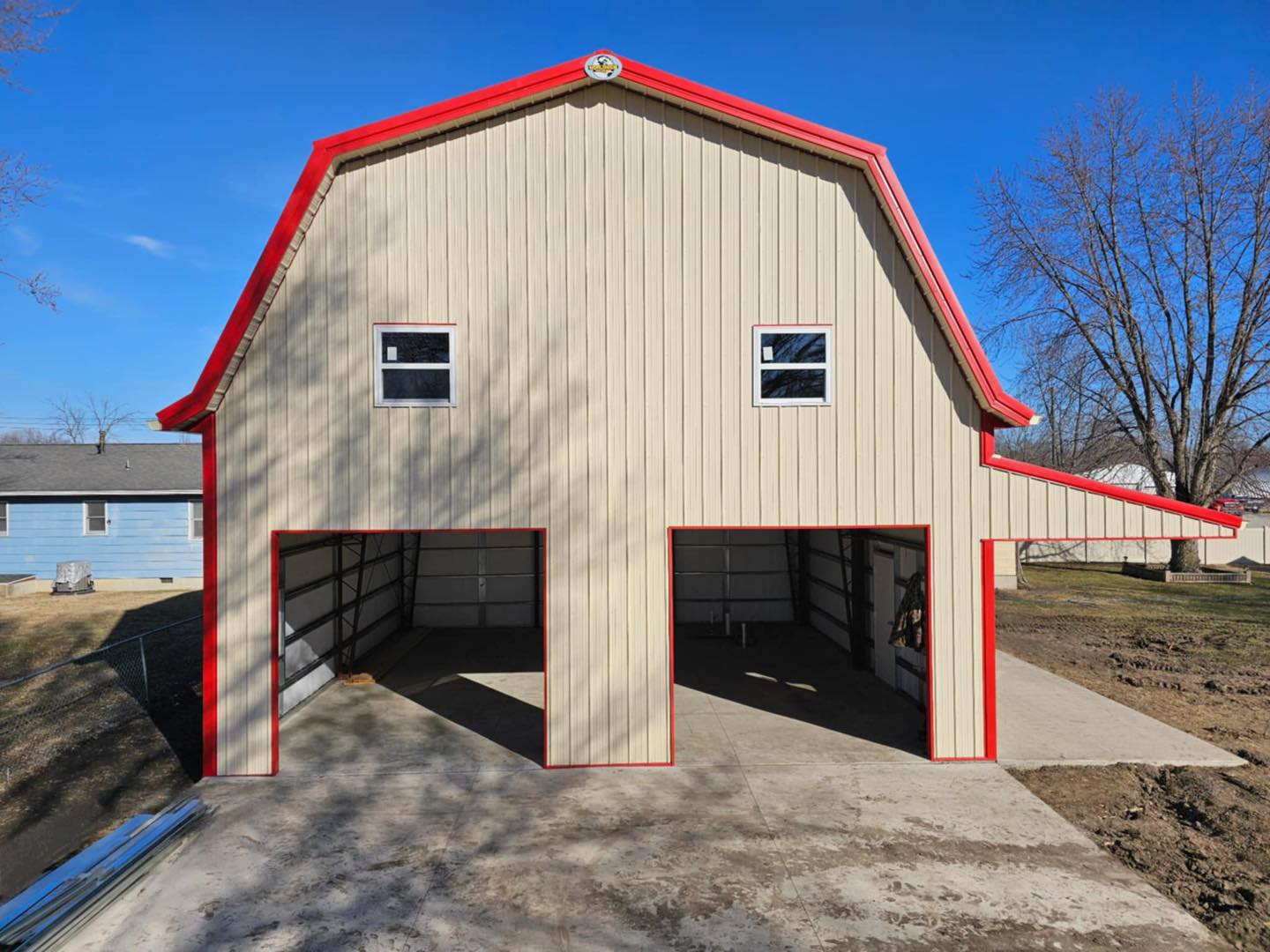 Beige two-story gambrel building with red roof, open garage doors; clear sky, trees, house in view.