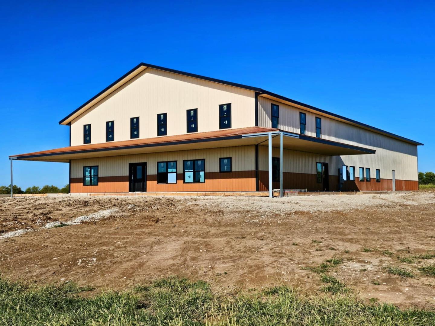 Two-story barndominium with brown/beige exterior, many windows, porch, and copper accents under blue sky.
