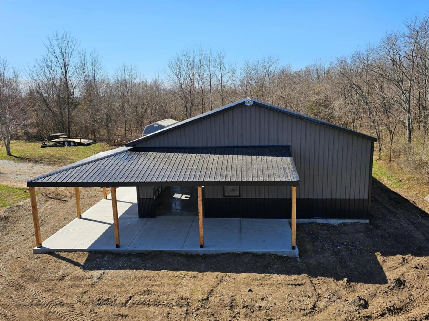 Aerial view of Litchford Garage: metal structure, black roof, porch, field, blue sky, trailer nearby.