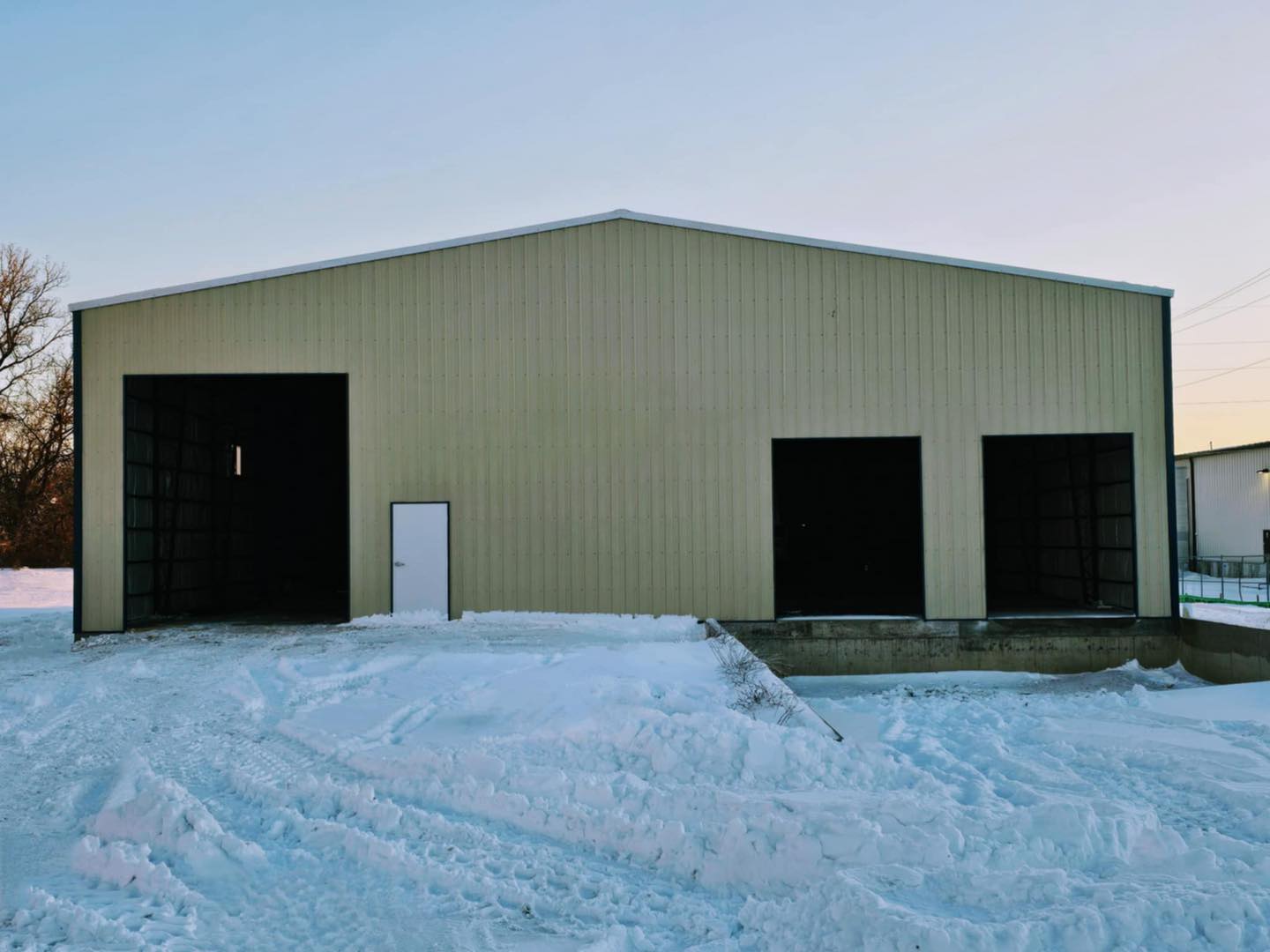 A large, beige industrial warehouse, home to ACCI Specialty Materials, stands on a snowy ground under a clear sky with three open garage doors. A small white pedestrian door is nestled between the larger openings. Sparse trees are visible to the left.