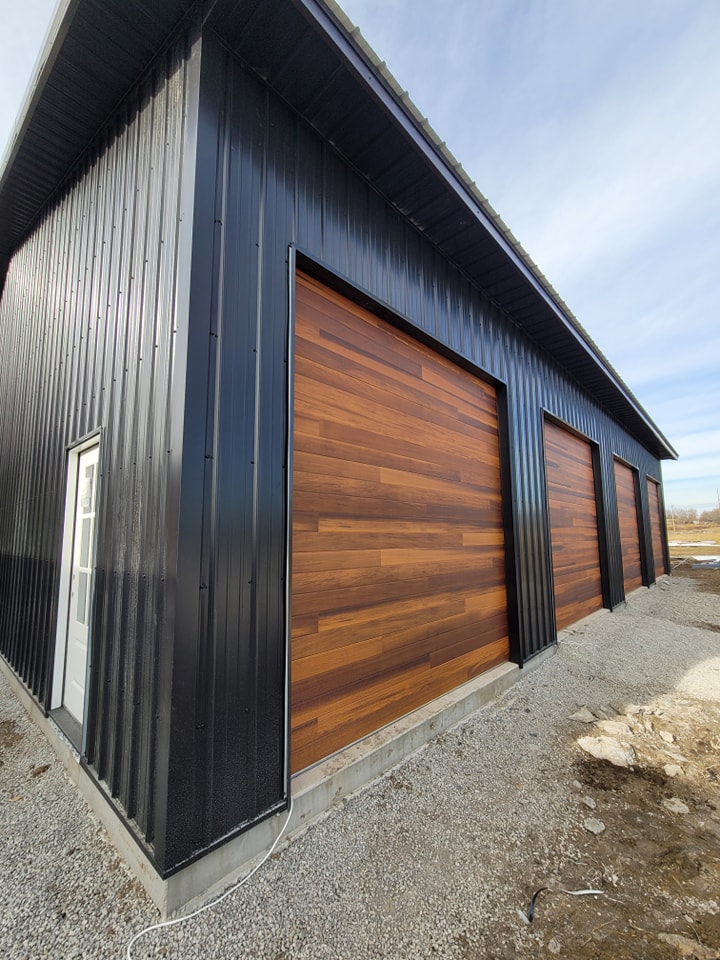 Modern building with three wooden garage doors, black siding, and white side door on a gravel driveway.