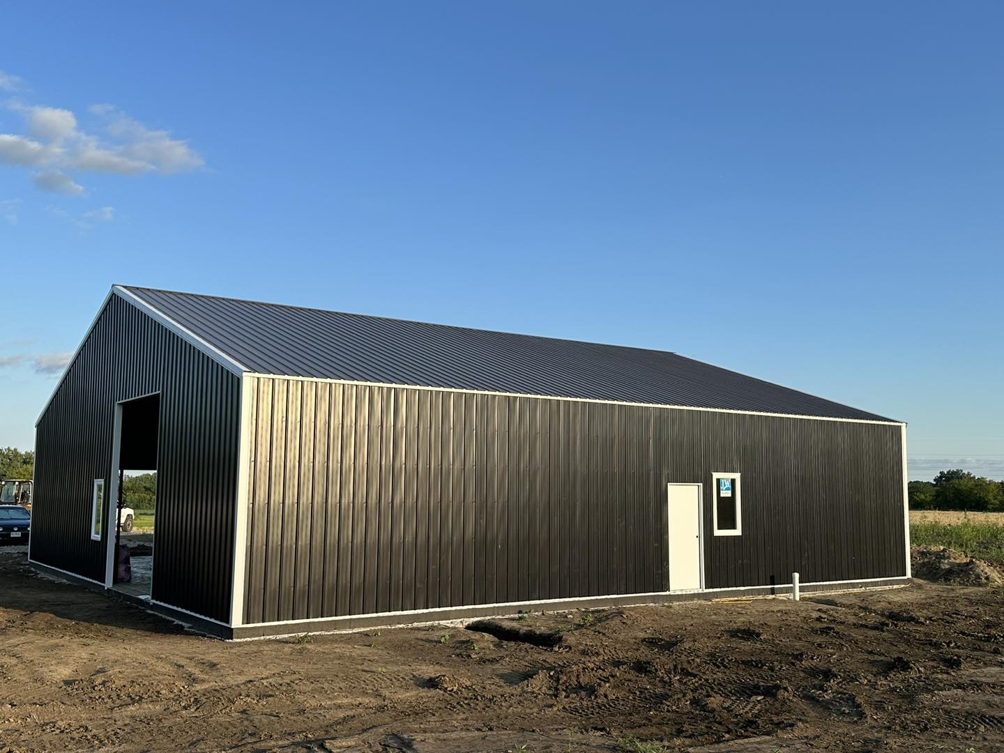 Barndominium: dark exterior, sloped roof, white door, small window; dirt ground; blue sky backdrop.