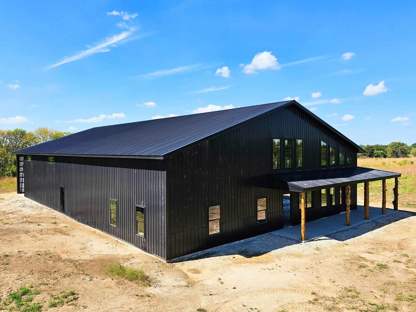 Black barndominium with metal roof and porch in open field; clear sky and sparse greenery.