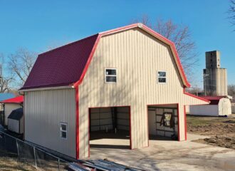 Beige barn with red roof, gambrel style, open doors, windows; silo and trees against blue sky.