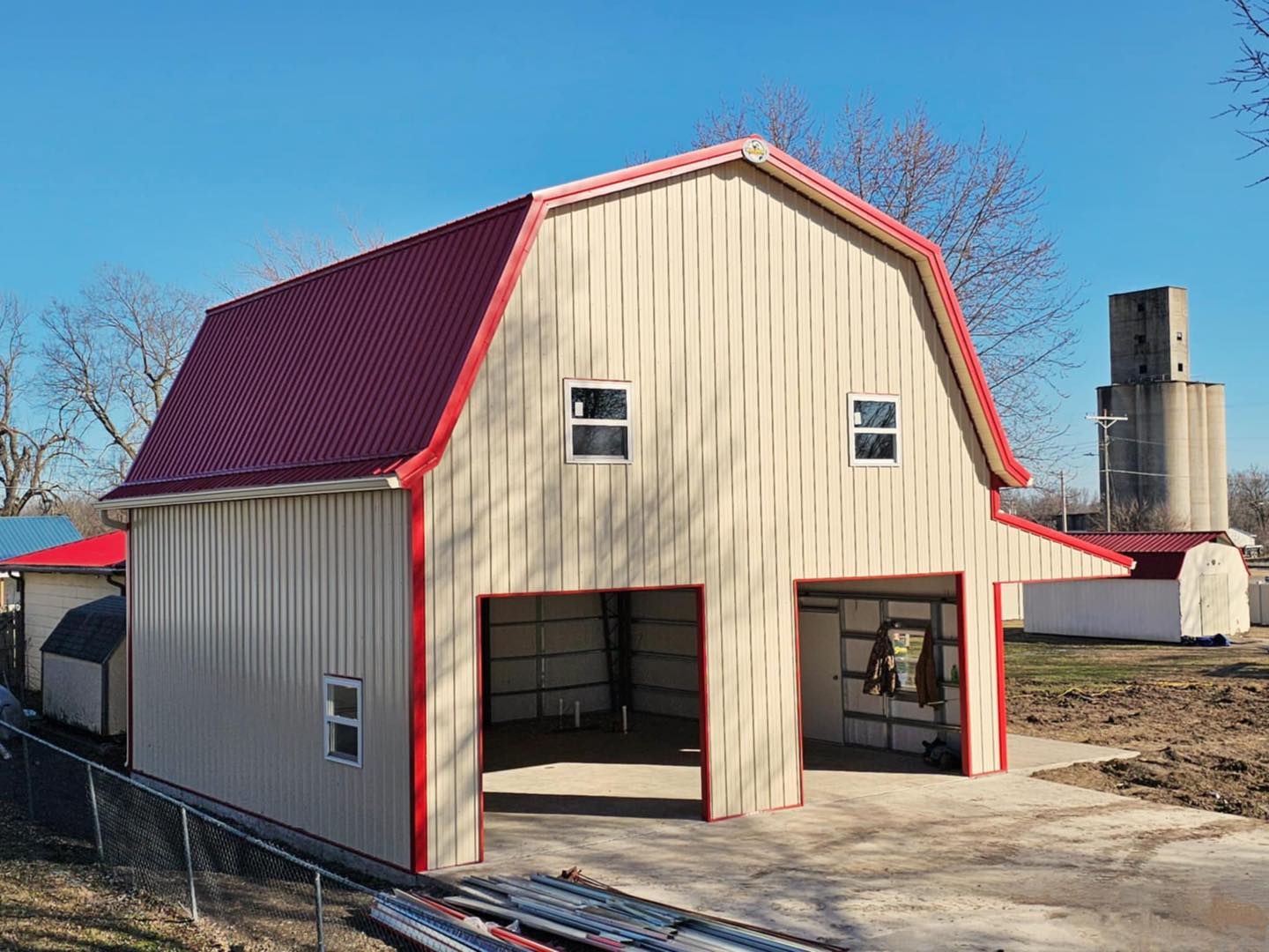 Beige barn with red roof, gambrel style, open doors, windows; silo and trees against blue sky.