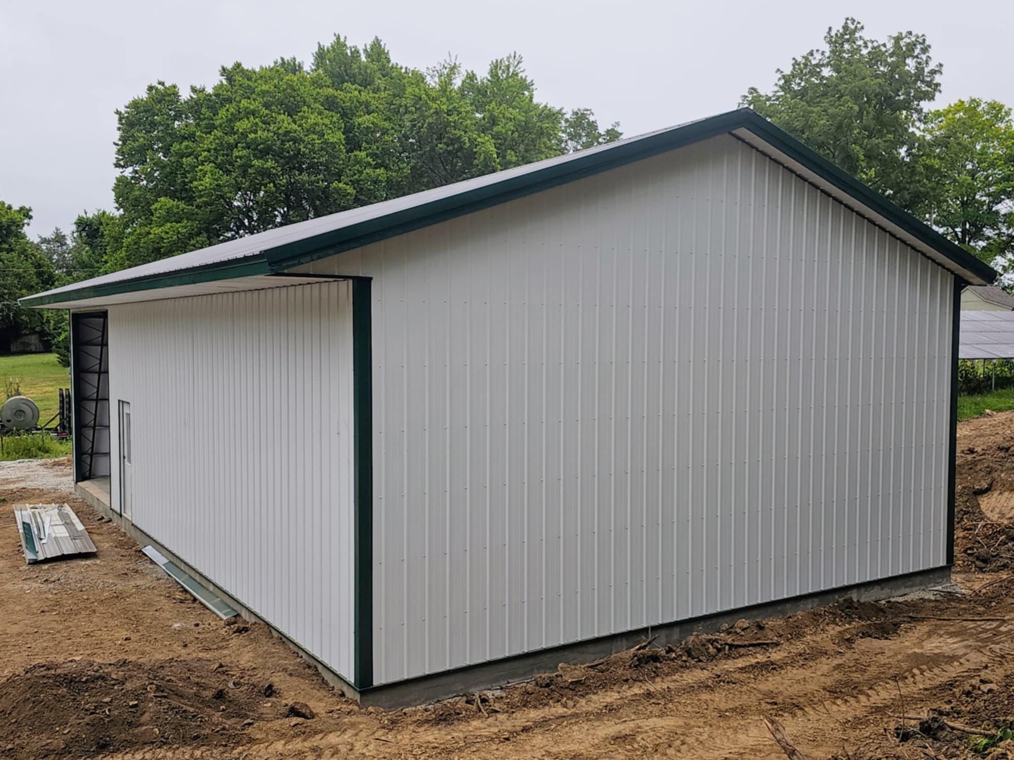 A large white metal shed with green trim by ChapCo, on a dirt area with trees and building materials nearby.
