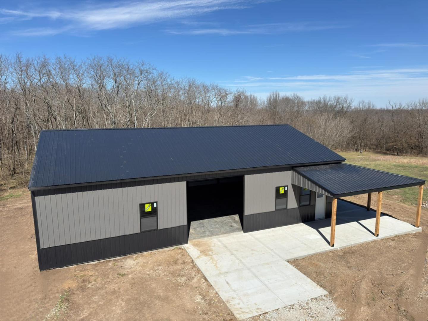 Aerial view of a metal barn with porch on concrete, amid bare trees and open land under clear sky.