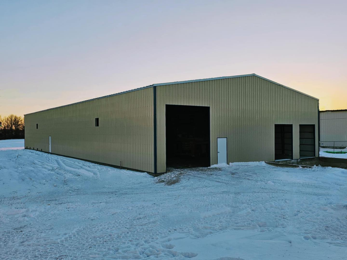 A large metal warehouse with an open entrance, surrounded by snow, stands in a flat, snowy landscape under a clear sky. The beige structure is constructed using specialty materials from ACCI, enhancing its durability and resilience against the harsh elements.