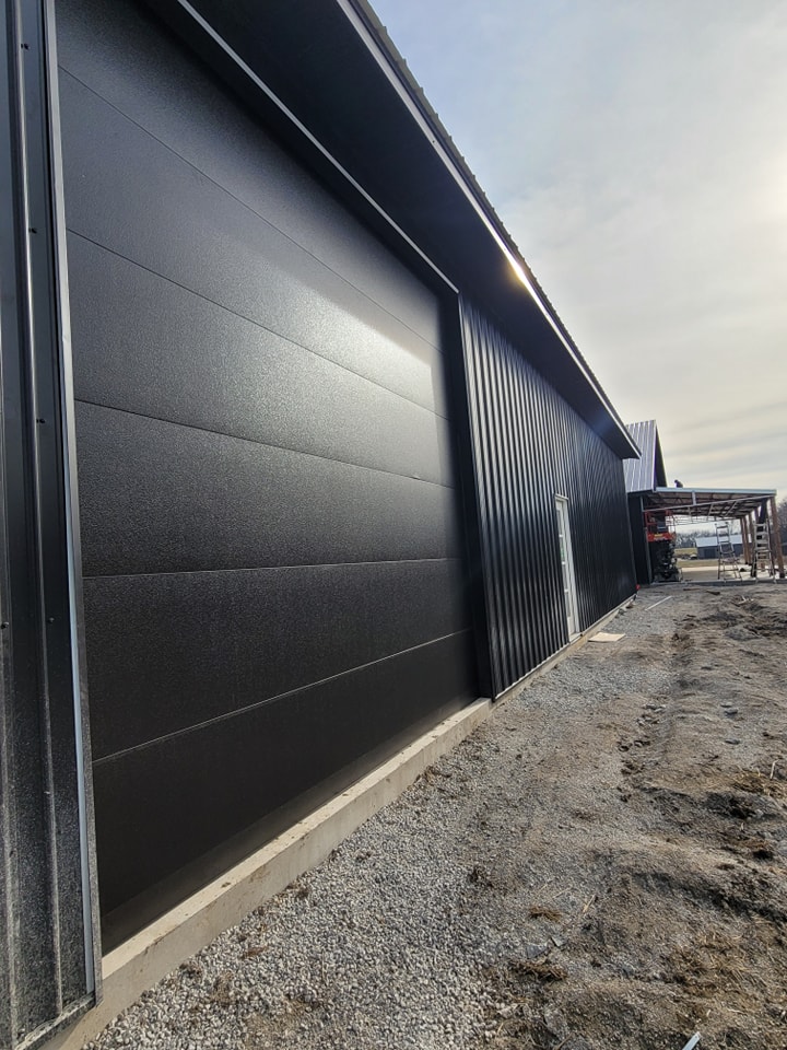 Industrial black building with large sliding door, gravel ground, and distant residential structure.