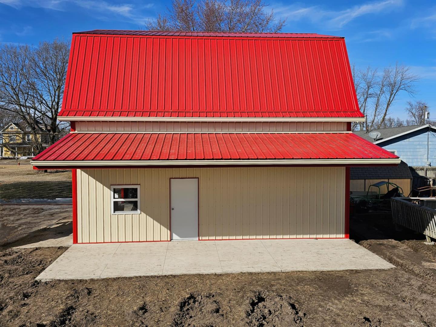 New beige barn with red roof, red-trimmed window/door, on concrete. Leafless trees and house nearby.