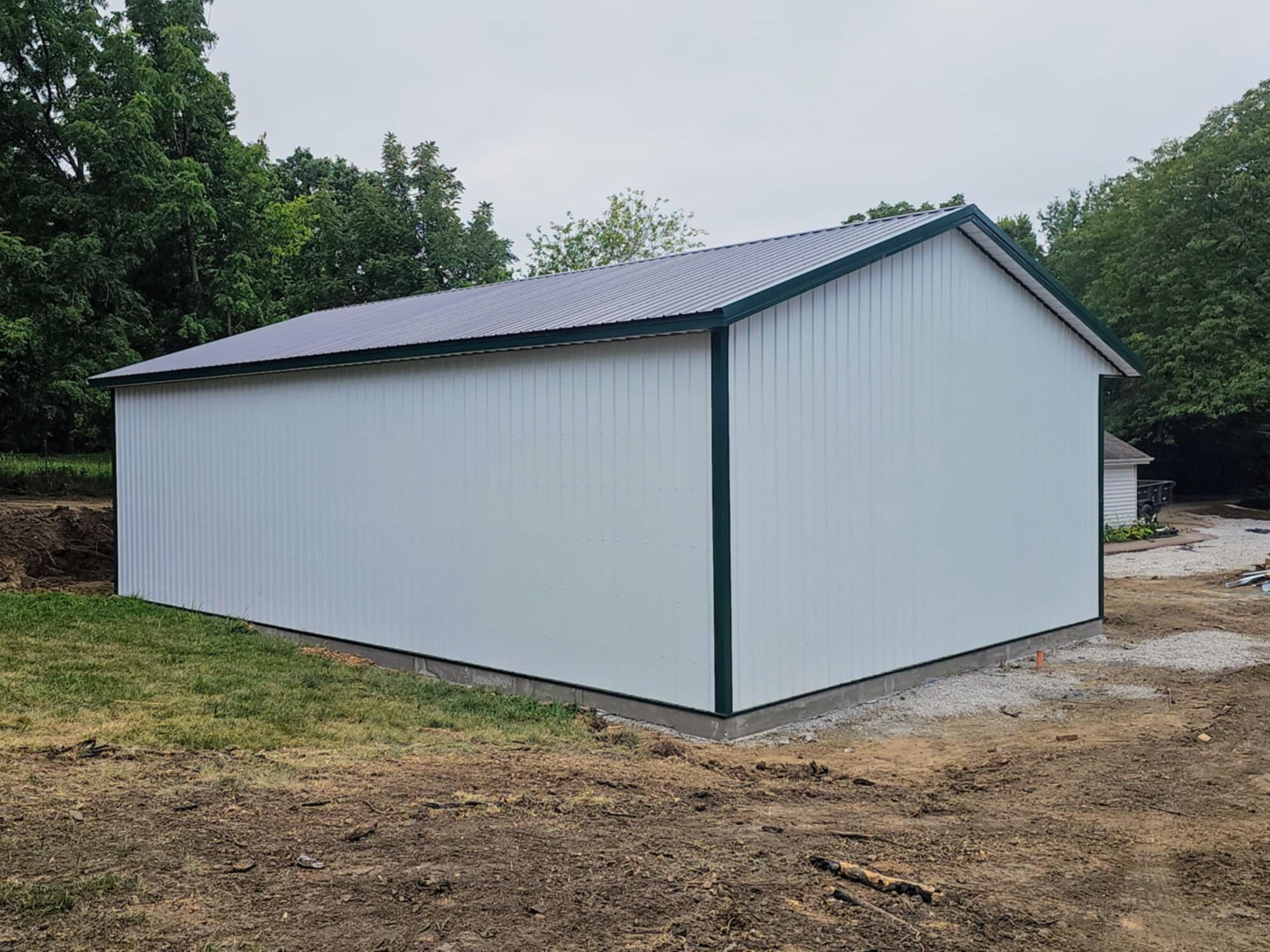New white metal building with green roof on concrete, surrounded by grass and trees under cloudy sky.