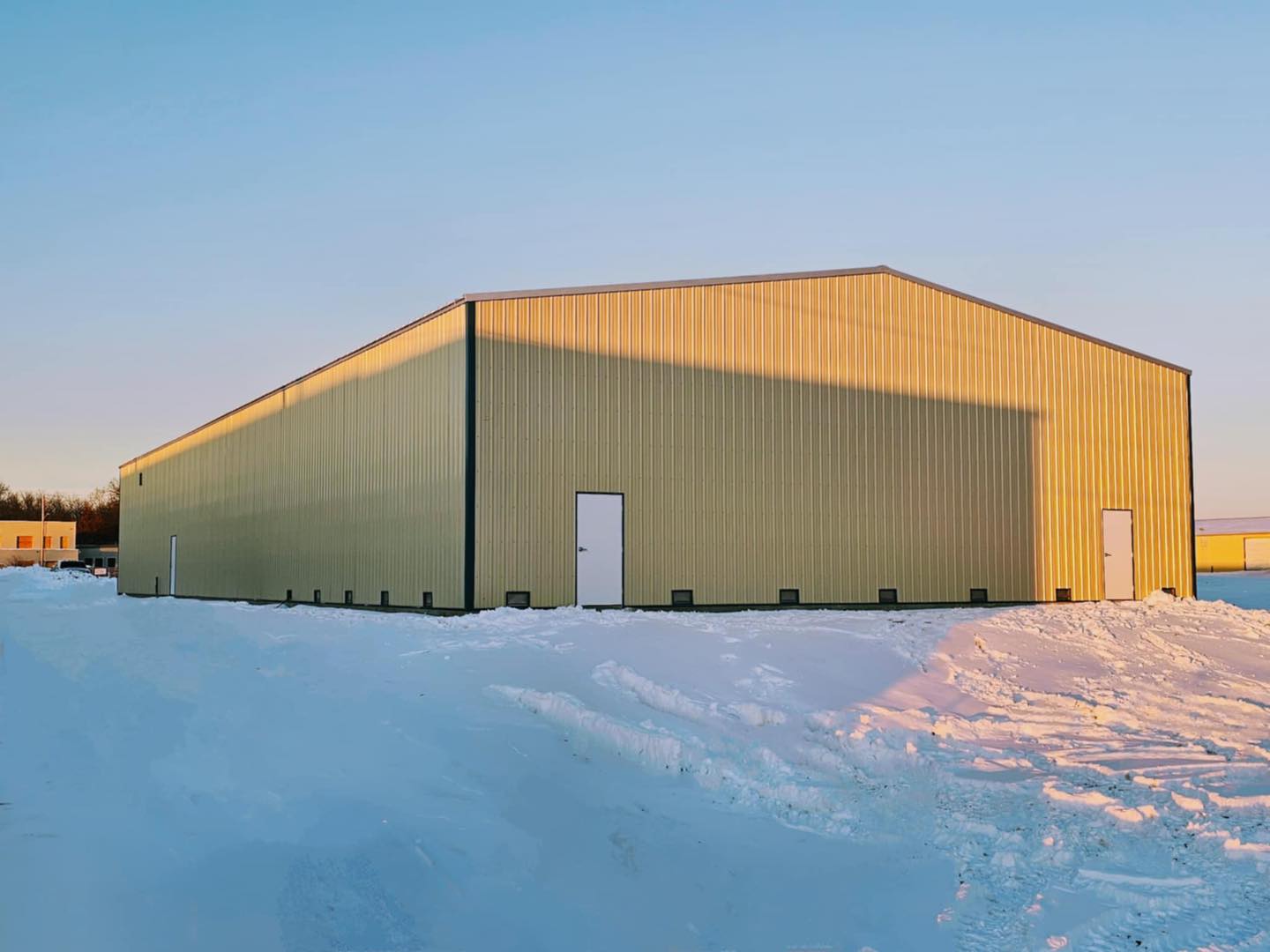 A large yellow metal building, rumored to store ACCI Specialty Materials, stands on a snowy landscape under a clear blue sky. The sun casts long shadows across the snow, while a few small structures peek out in the distance.