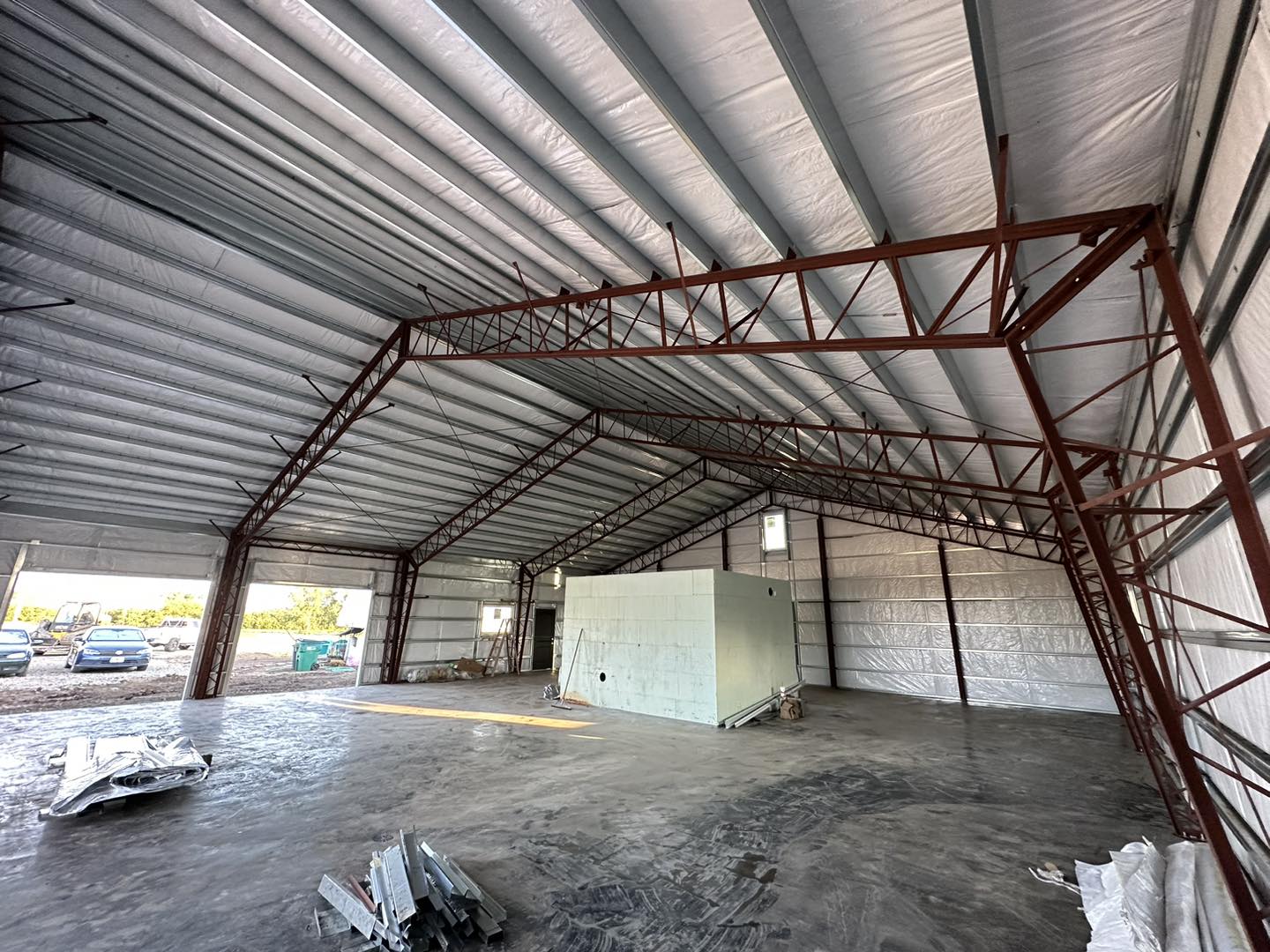 Unfinished warehouse interior with metal framework, large windows, and a view of cars and trees outside.