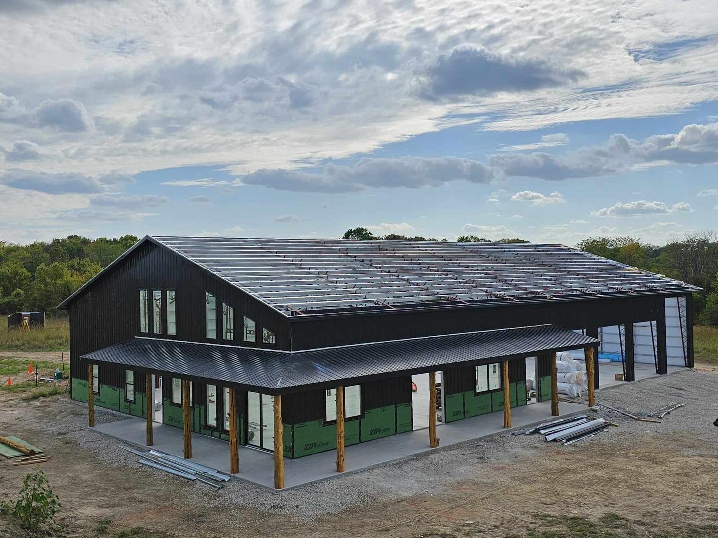Large black barndominium with metal roof on grass, under cloudy sky, surrounded by construction materials.