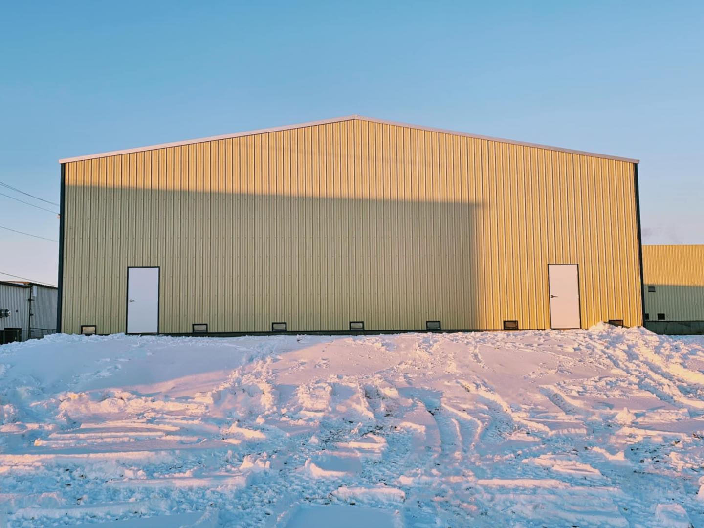 A large, yellow metal building with white doors stands on a snow-covered landscape under the clear blue sky. As the setting sun casts a shadow on its facade, it highlights the distinct design of ACCI Specialty Materials' innovative architecture.