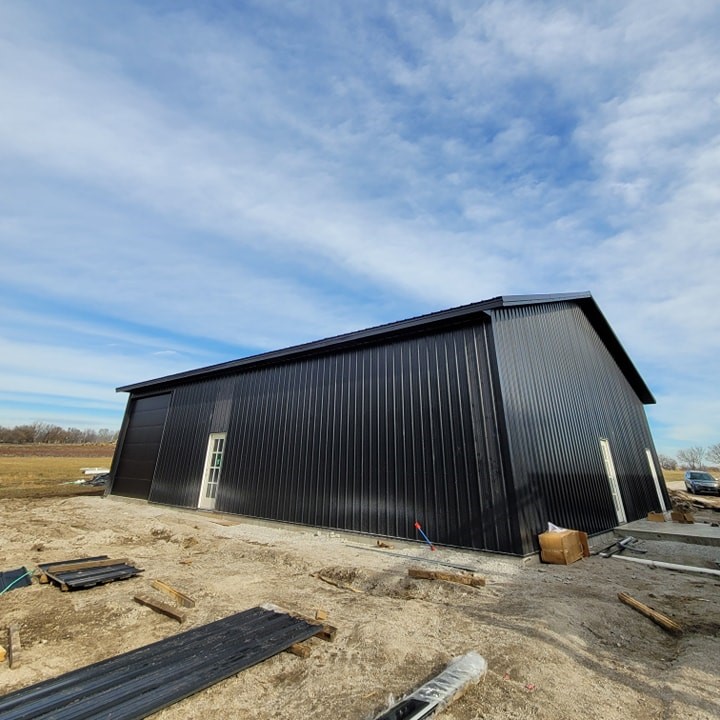 Black barn-style building with a sloped roof, construction materials around, open field in the background.