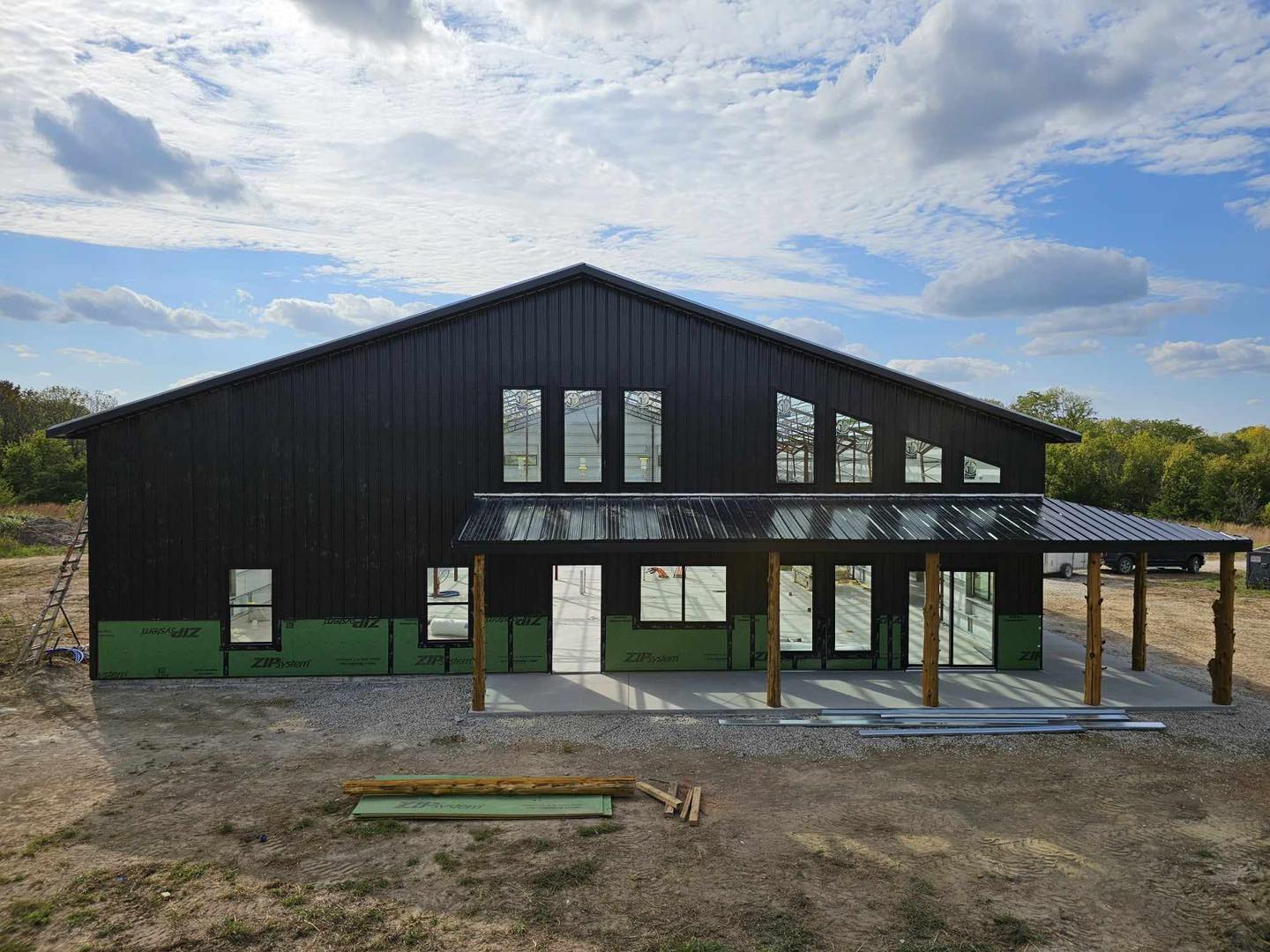 Dark barndominium with metal roof, windows, porch, and scattered materials on grassy area under clouds.