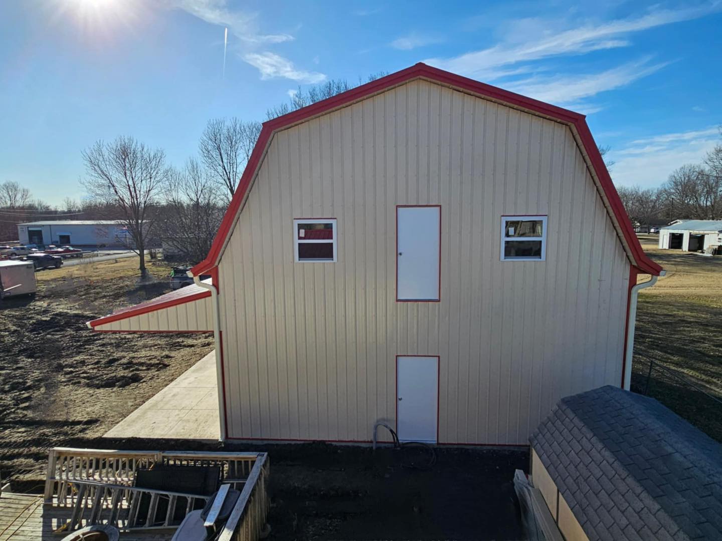 Beige barn with red roof in rural setting, clear sky, leafless trees, small shed in background.