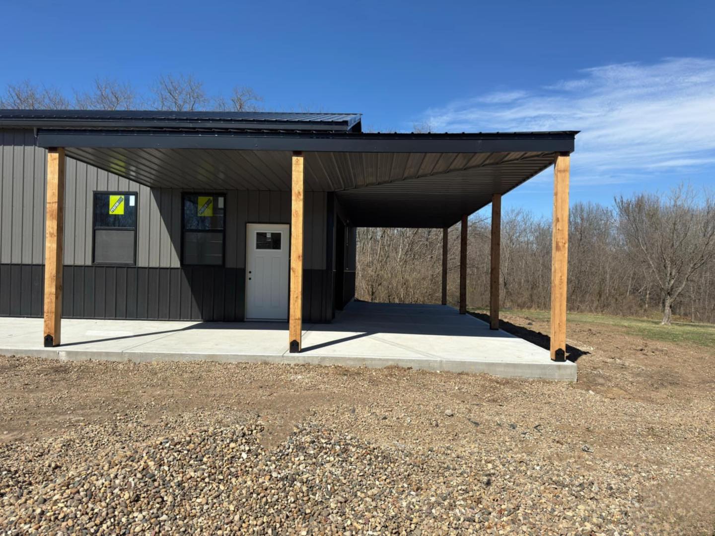 Modern building with wood, metal, porch on gravel lot; leafless trees & clear sky in background.