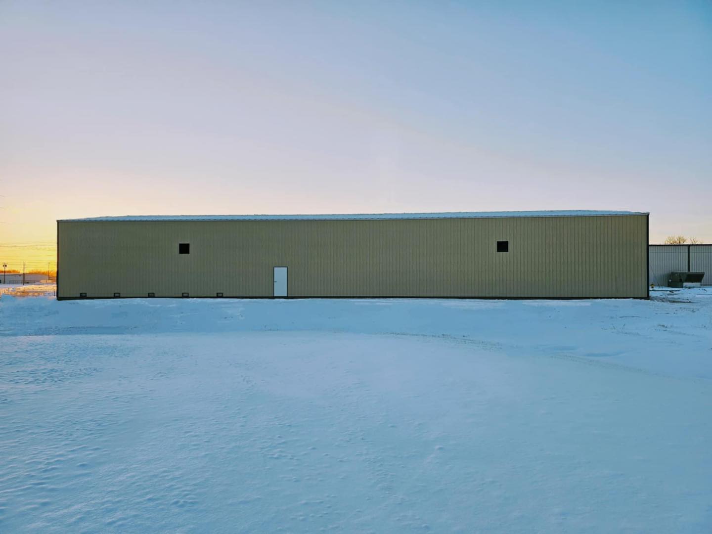 A large, rectangular beige warehouse from ACCI Specialty Materials stands alone on a snow-covered landscape under a clear blue sky at sunset. There are a few small windows and a single white door, with a hint of another building visible on the right side.