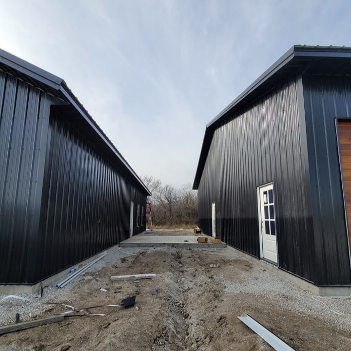 Two black metal buildings with white doors stand parallel on gravel, leading to trees and cloudy sky.