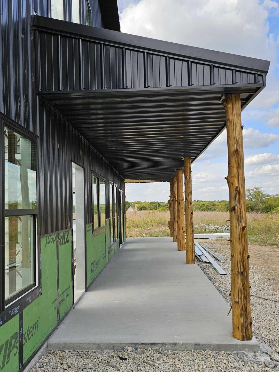 Under a cloudy sky, a black barndominium with metal siding and wooden beams stands in a grassy field.