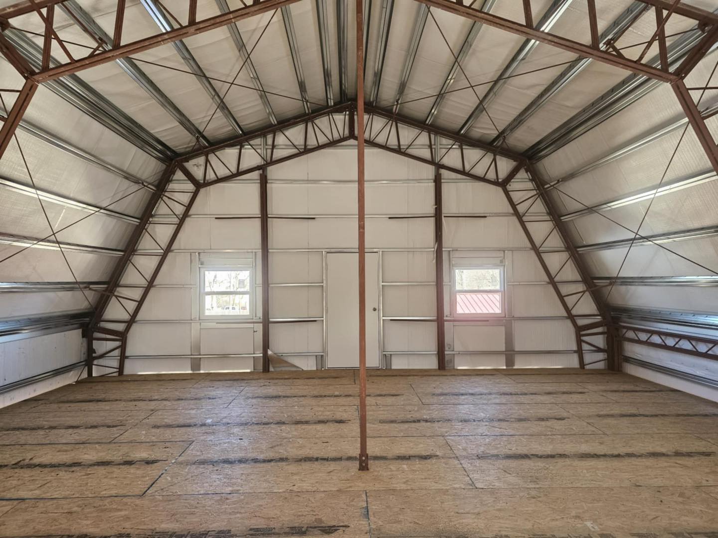 Unfinished metal interior with steel beams, plywood floor, two windows, and natural lighting.