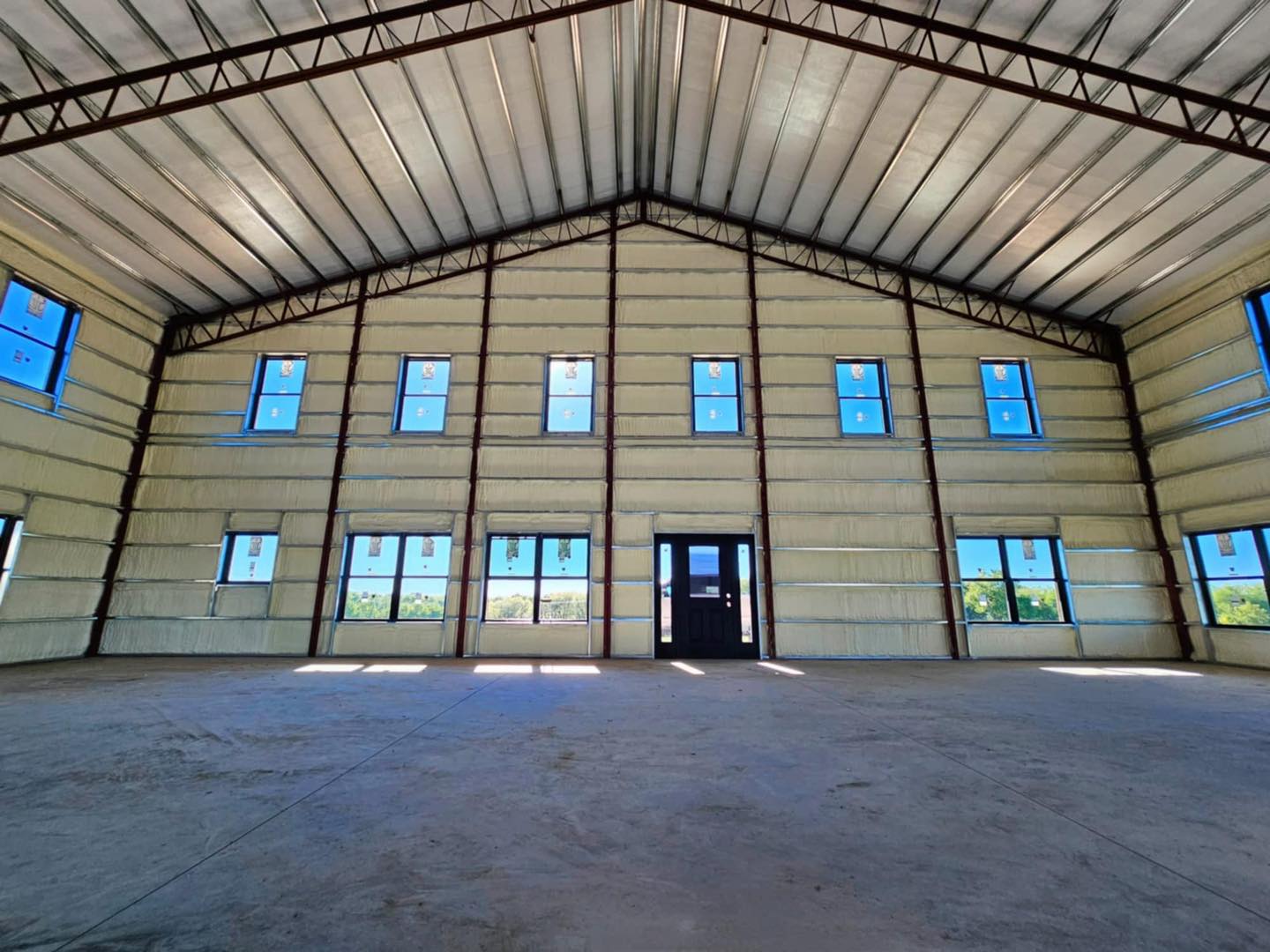 Interior of a spacious metal warehouse with high ceilings, windows, and central doors, lit by sunlight.
