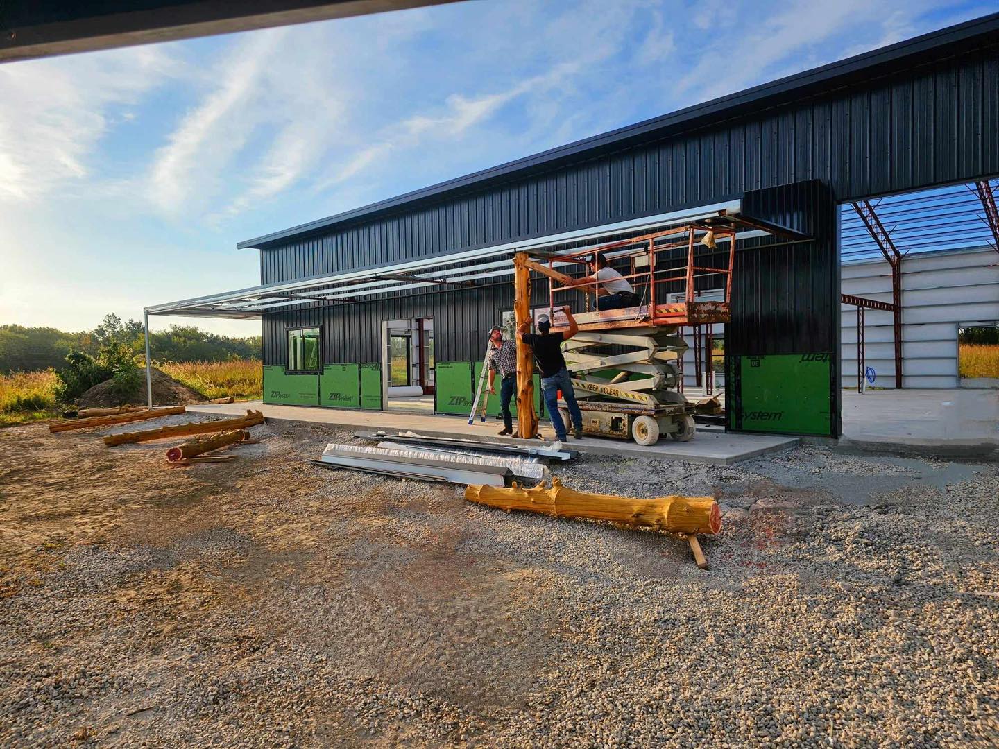Workers use a lift to install black metal panels on a barndominium-like building's steel frame.