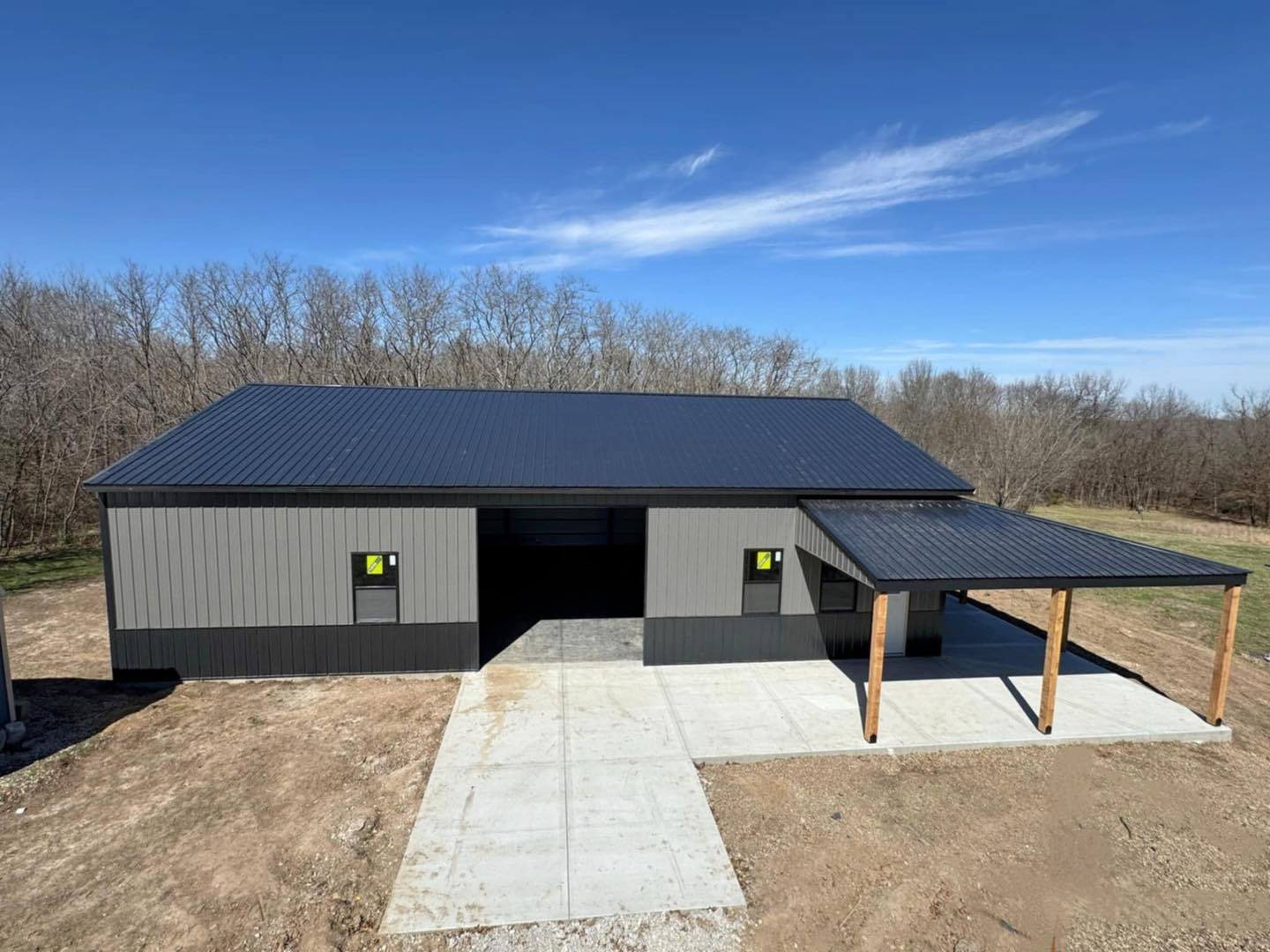 Gray metal garage with pitched roof and covered area, set on an open plot with bare trees and blue sky.