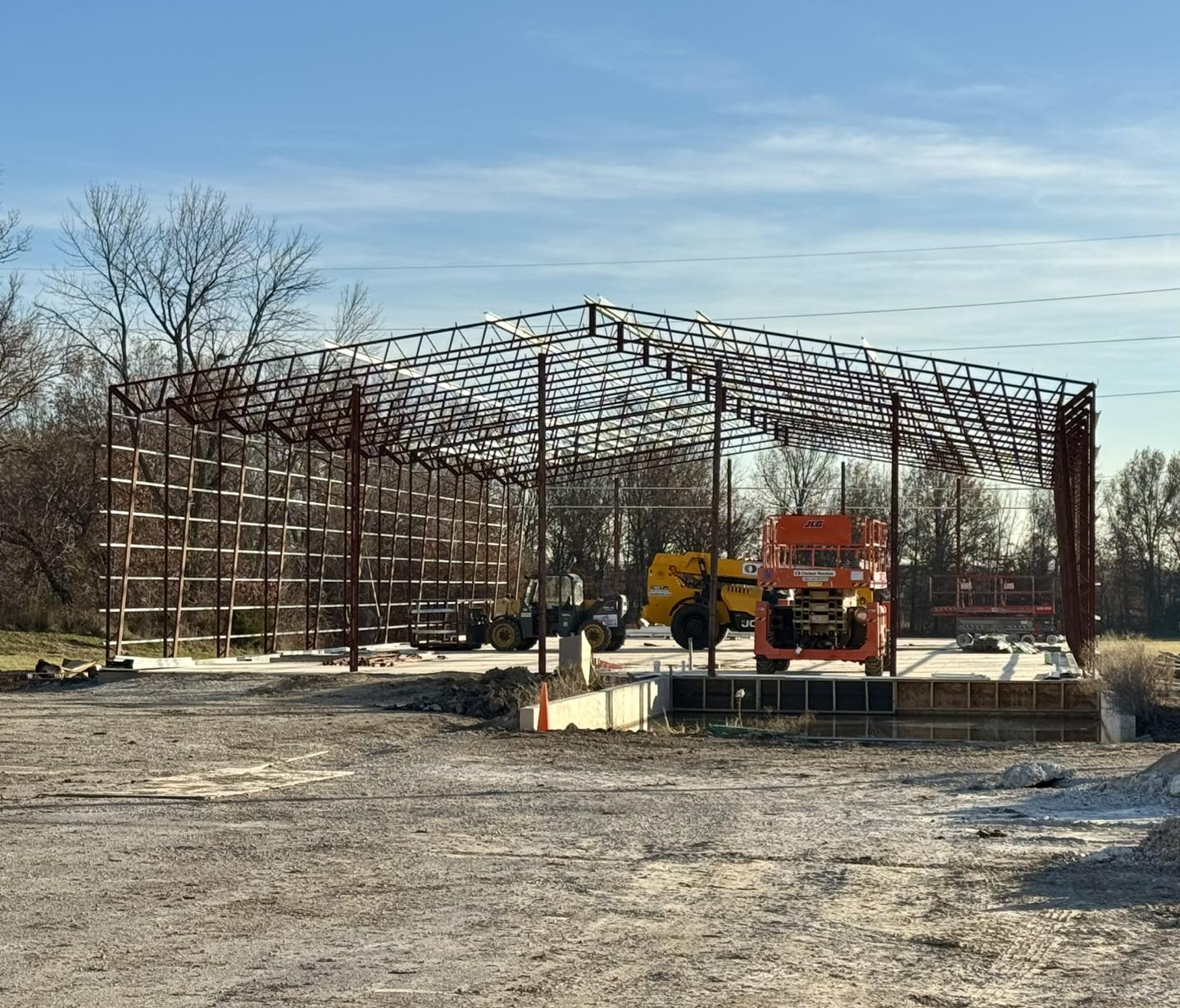 A construction site showcases a metal framework of a large building in progress, utilizing specialty materials. Various construction vehicles and equipment, including an ACCI scissor lift and a forklift, are present. Trees and a clear blue sky form the backdrop.