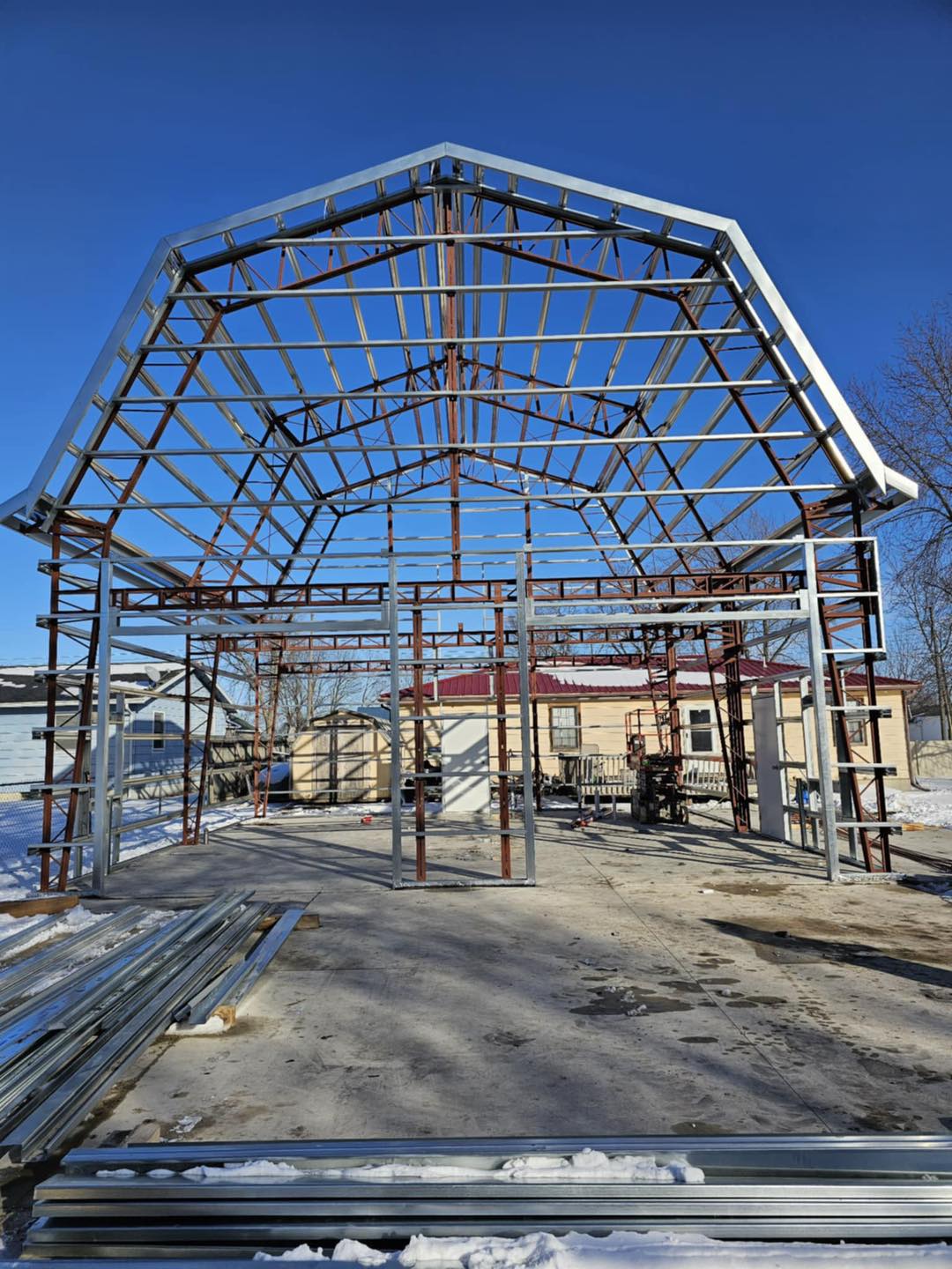 Metal frame on concrete foundation, blue sky visible, with trees and houses behind; beams nearby.