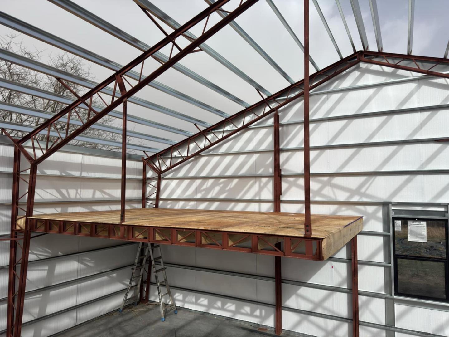 Building interior under construction with metal framework, wooden platform, ladder, and shadow patterns.