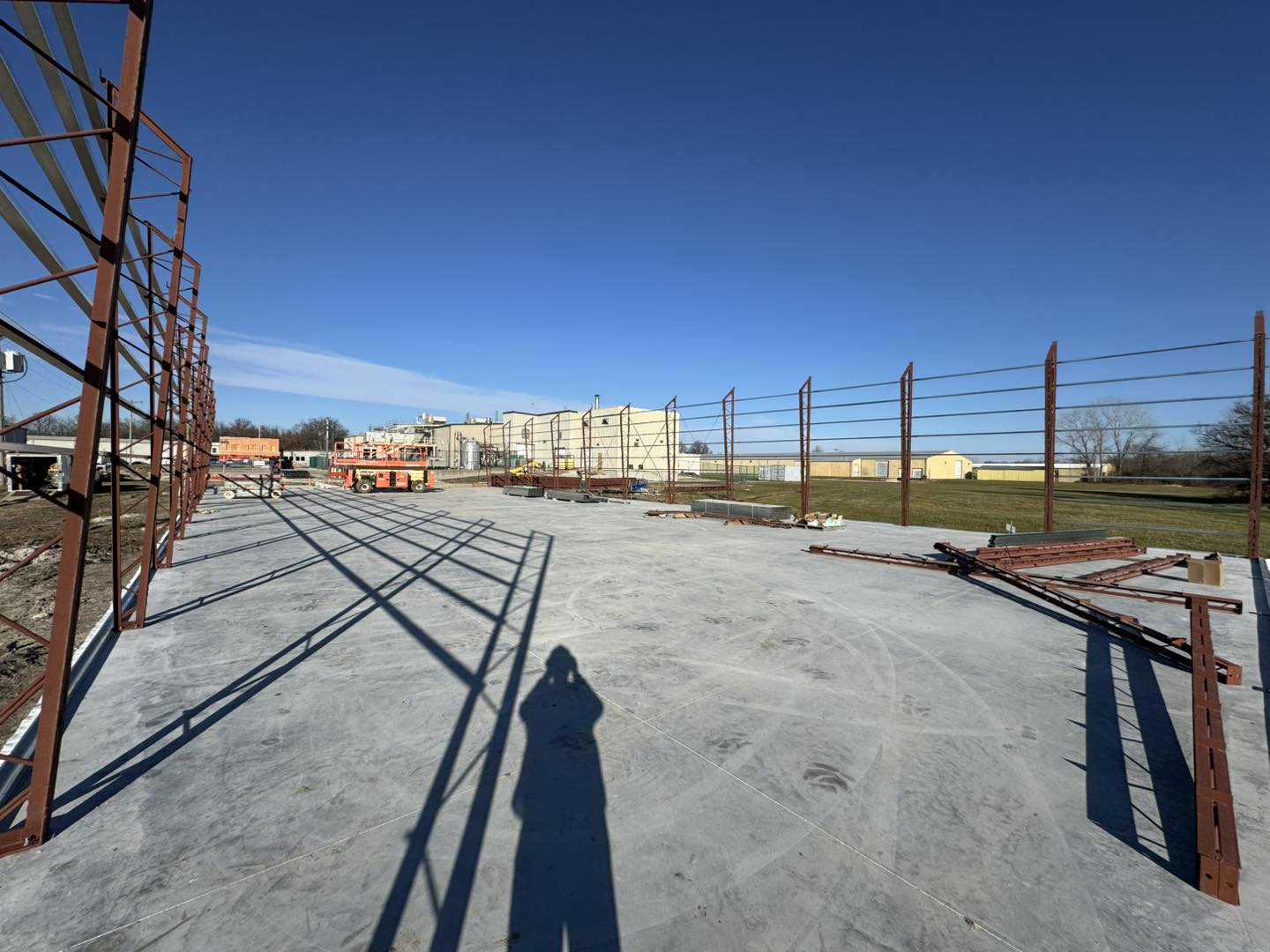 Under a clear blue sky at the construction site, steel frames rise from a concrete foundation, showcasing ACCI's specialty materials. Construction equipment and scattered supplies create an industrial tableau, as the shadow of a worker stretches toward the large building in the background.