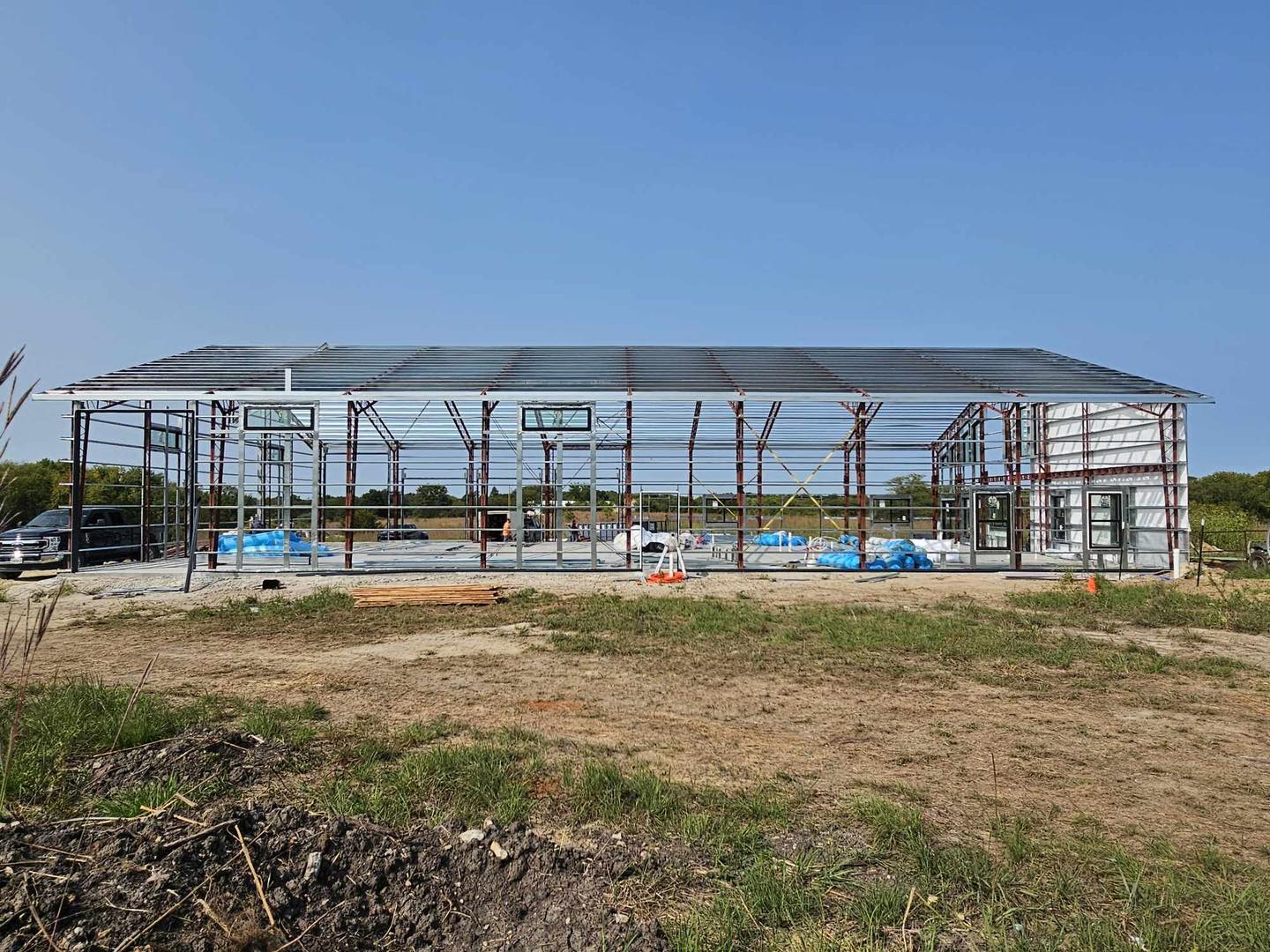 Metal-framed structure under construction, with roofing and panels, vehicles on grassy site.