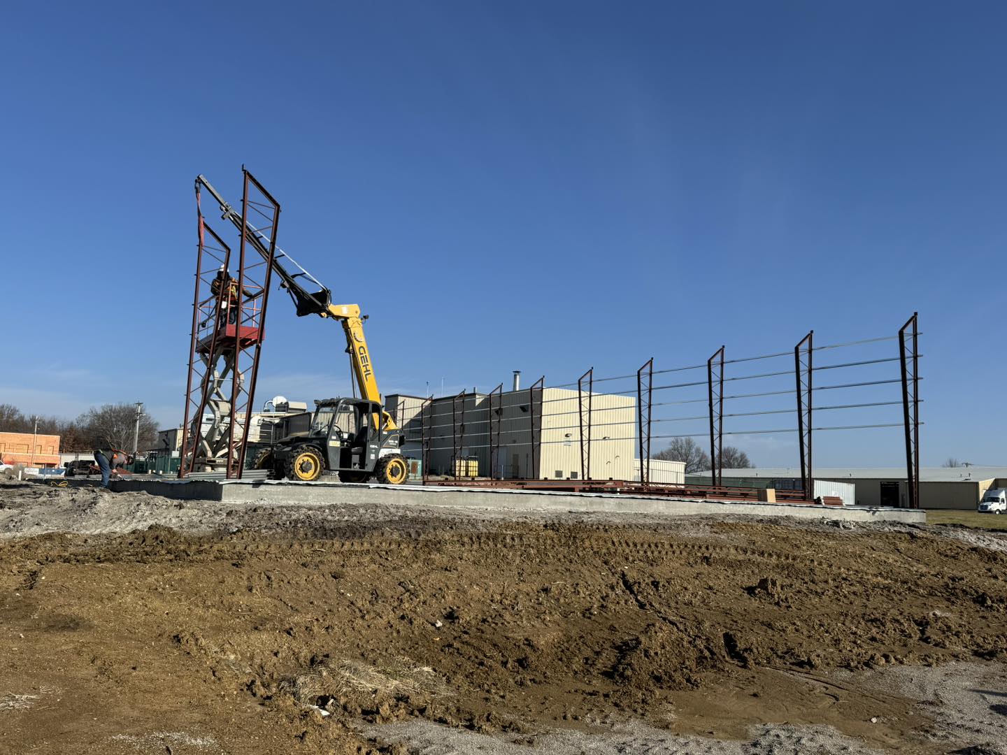 At the construction site, a yellow crane hoists a metal beam from ACCI to workers on a steel structure. The ground, scattered with specialty materials and dirt, leads to a large warehouse standing proudly under the clear blue sky.