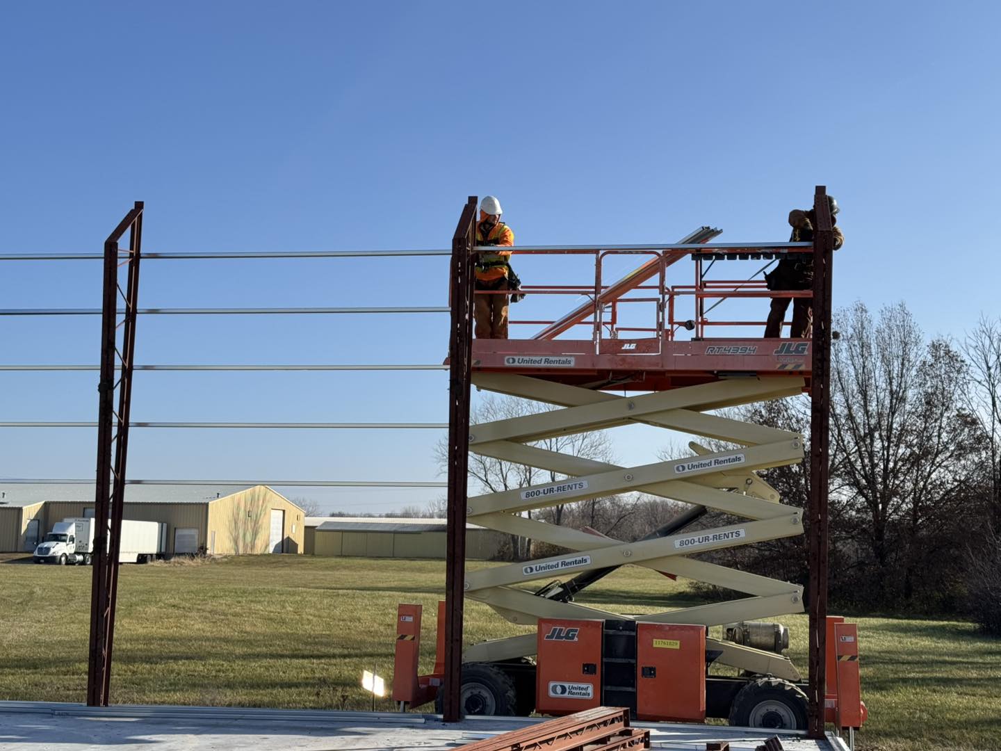 Two workers in safety gear are on an elevated platform, expertly handling specialty materials to install the metal framework for a building. The platform is extended between two steel supports, set against a clear day with grass and trees in the background.