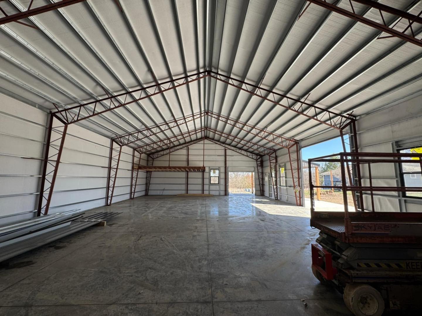 Unfinished warehouse with steel beams, metal roof, sunlight streaming through an open door, and a scissor lift.