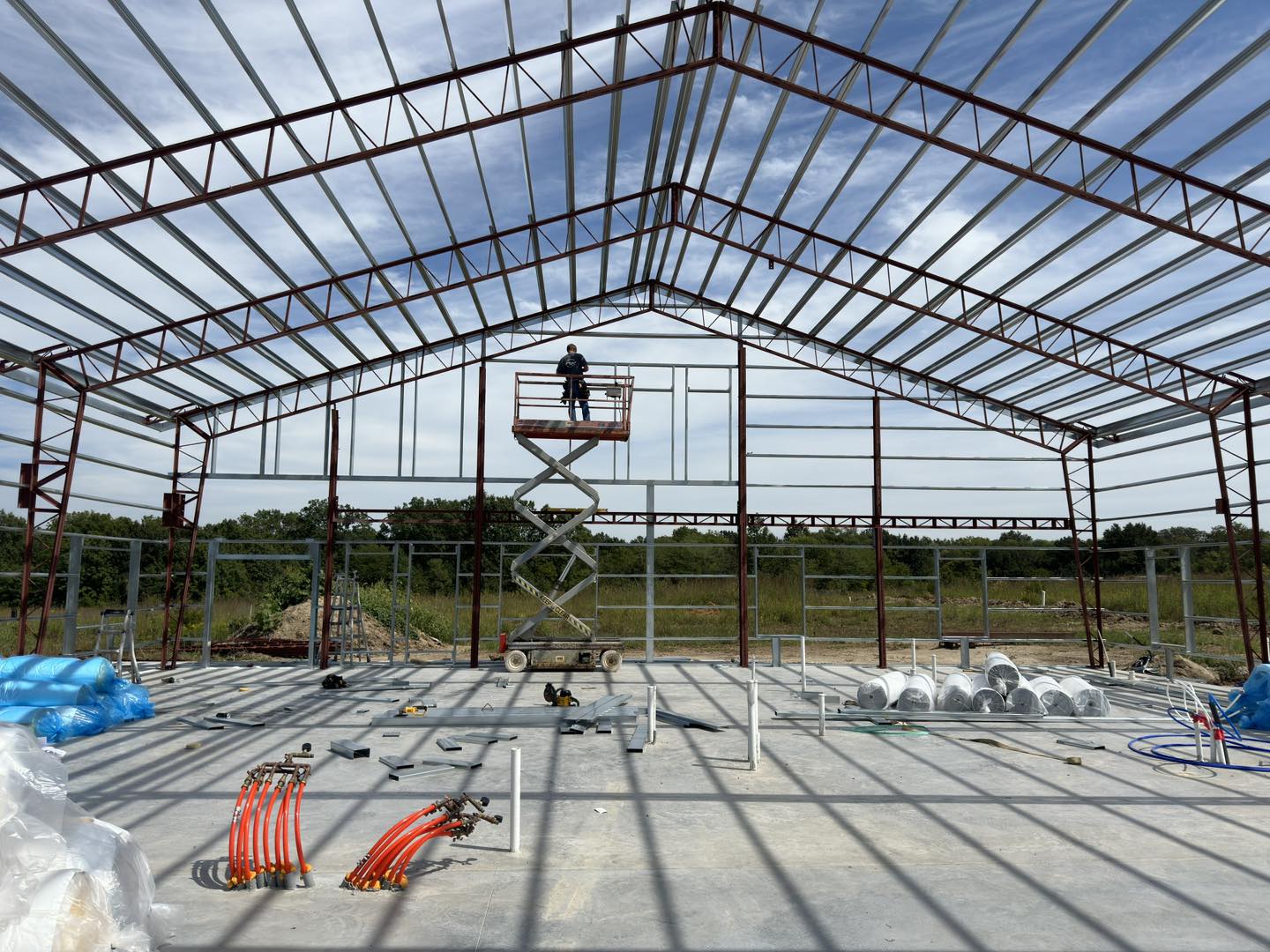 A worker on a lift under a black steel-frame barndominium, with cables and tools on the concrete floor.