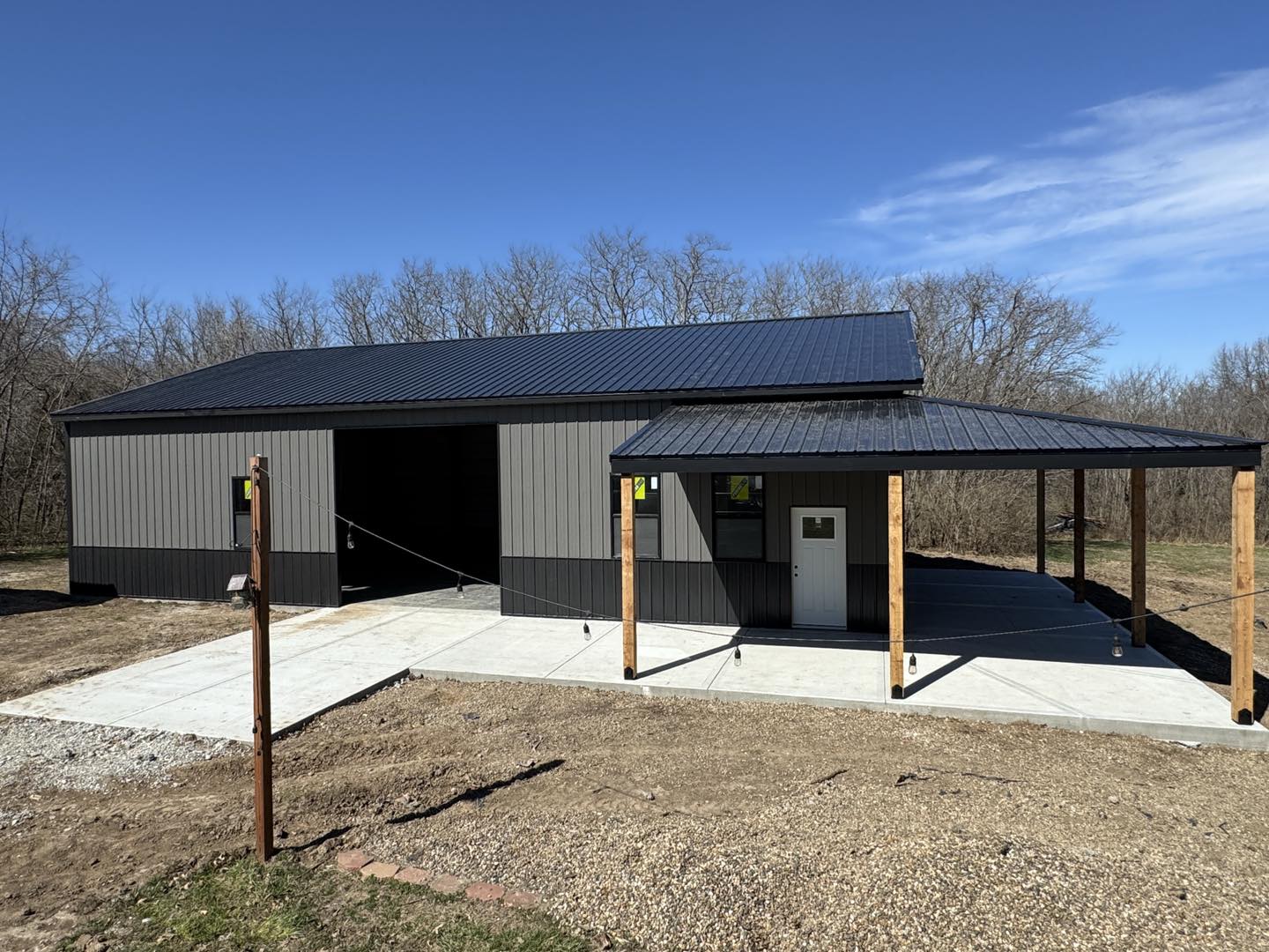 Metal building with sloped roof and carport, on concrete driveway, trees and blue sky in background.