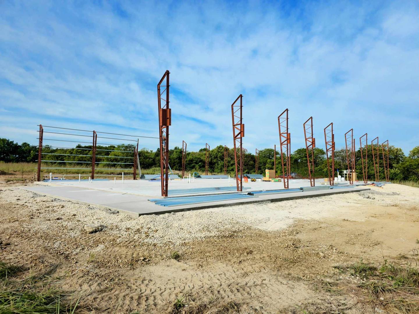 Construction site with concrete foundation and metal beams, set against trees and a blue sky.