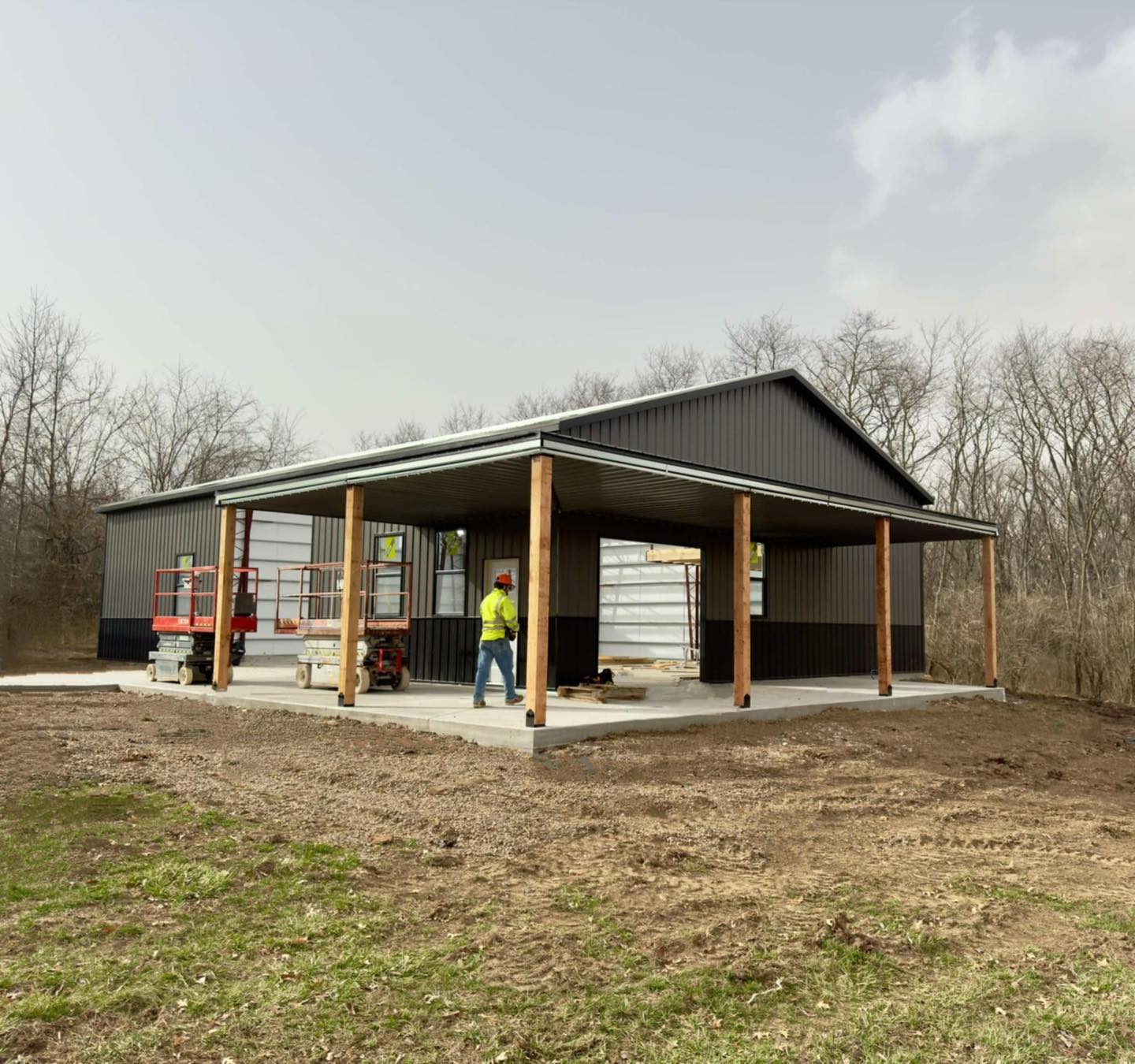 Metal garage under construction with a worker, scissor lift, and materials; trees in the background.