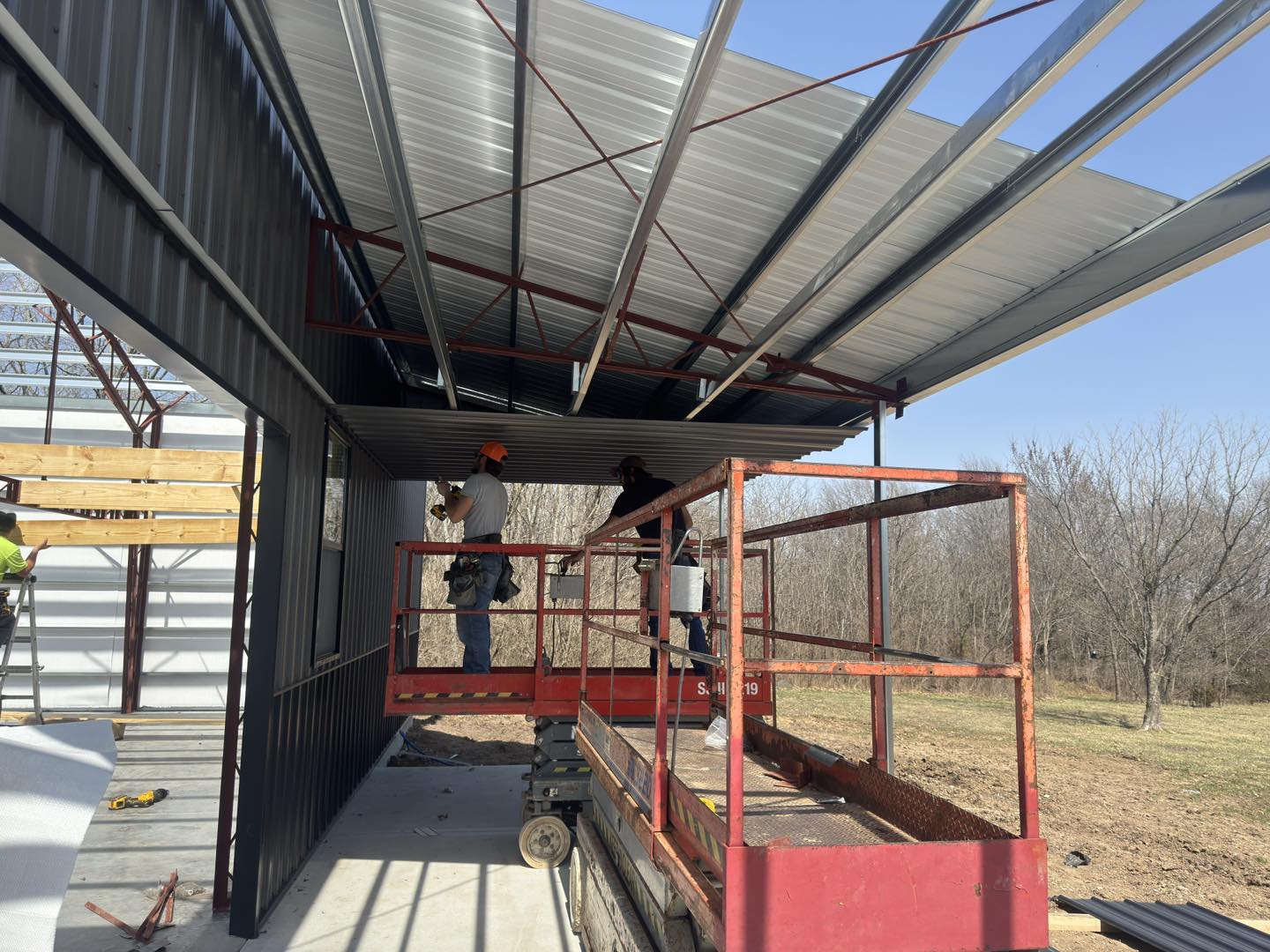 Two workers on a cherry picker install a roof on a metal garage, trees and clear sky in the background.