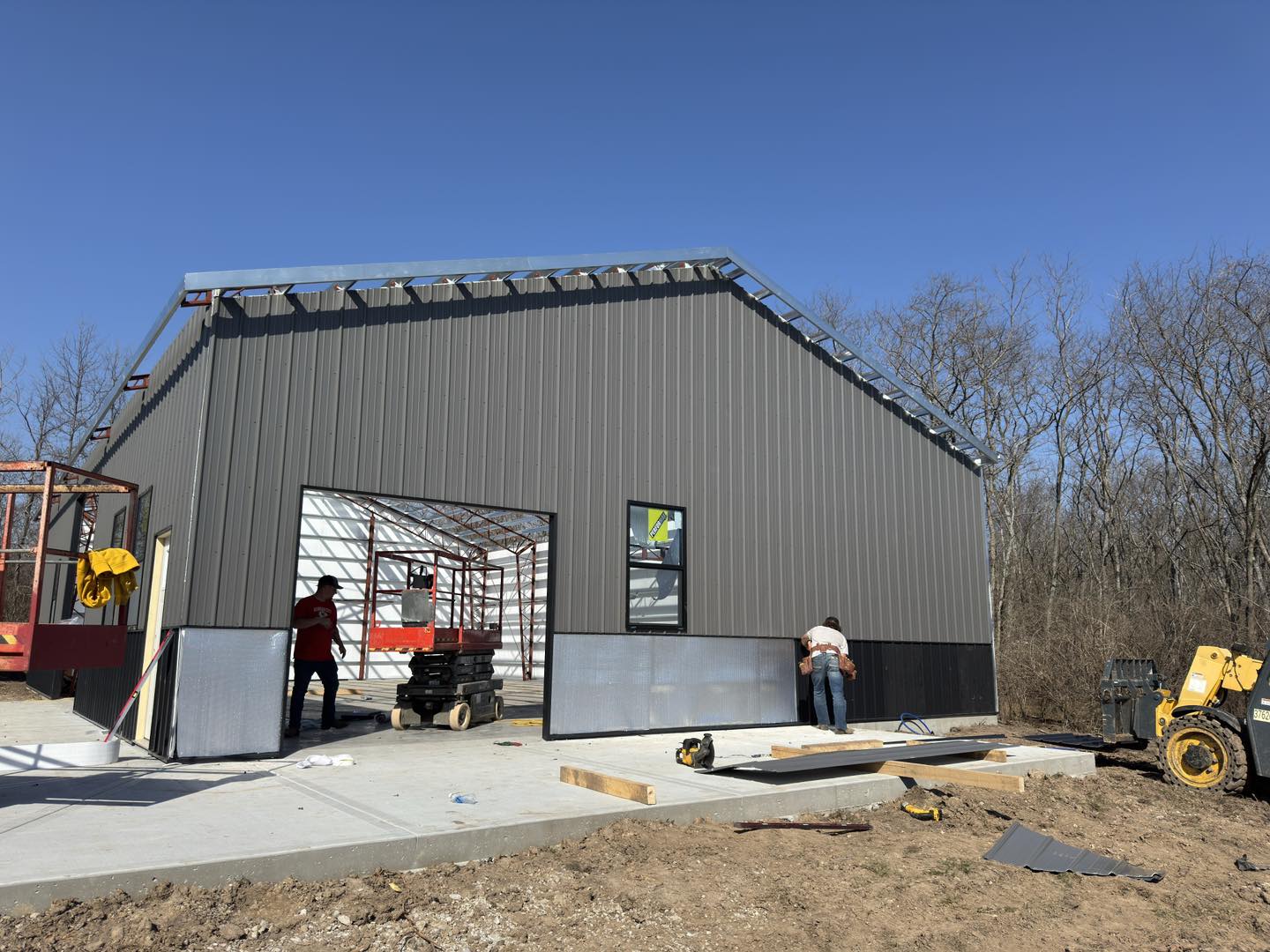 Workers install panels on a metal garage under clear skies, surrounded by leafless trees and equipment.
