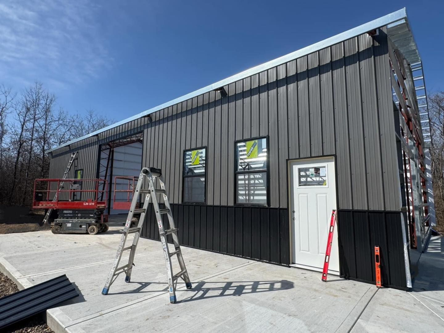Partially built metal shop with gray siding, large windows, ladder, and lift vehicle beside lush trees.