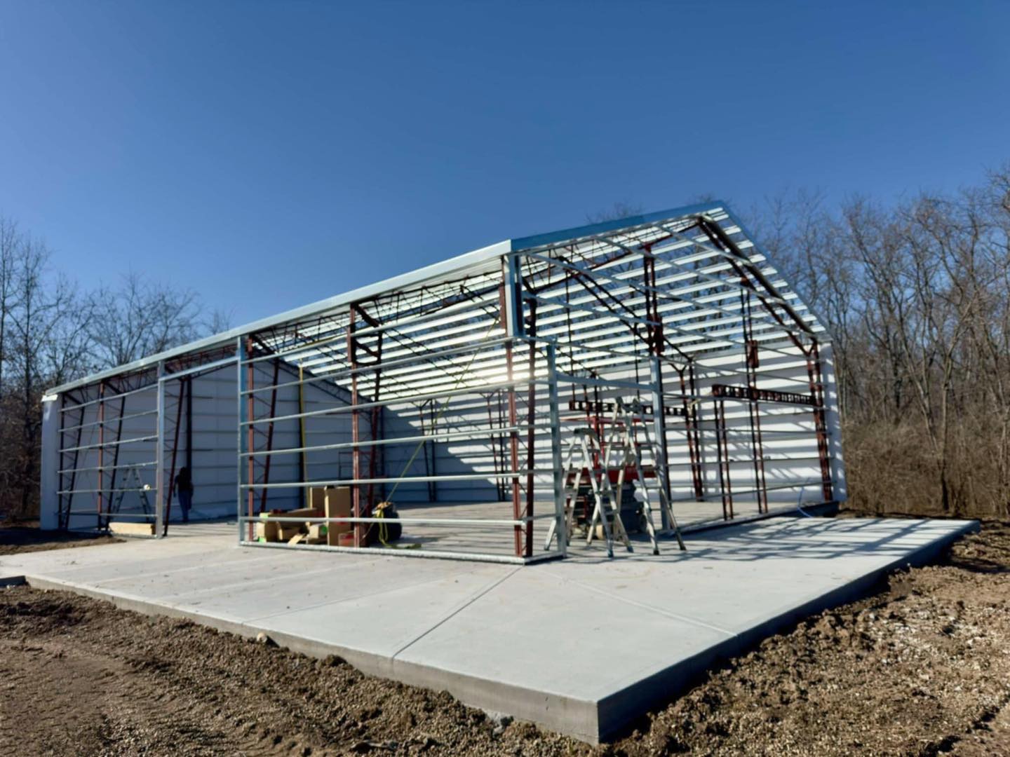 Partially built metal garage on concrete, with construction tools and trees under a clear blue sky.