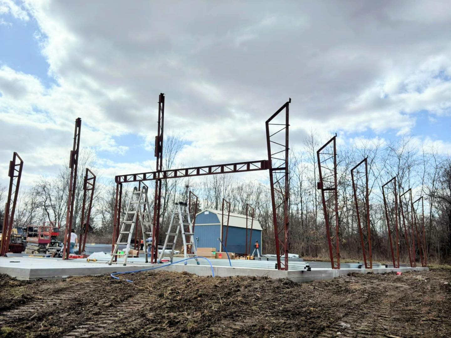 Partially built metal framework on concrete, ladders around, blue building and trees in cloudy background.