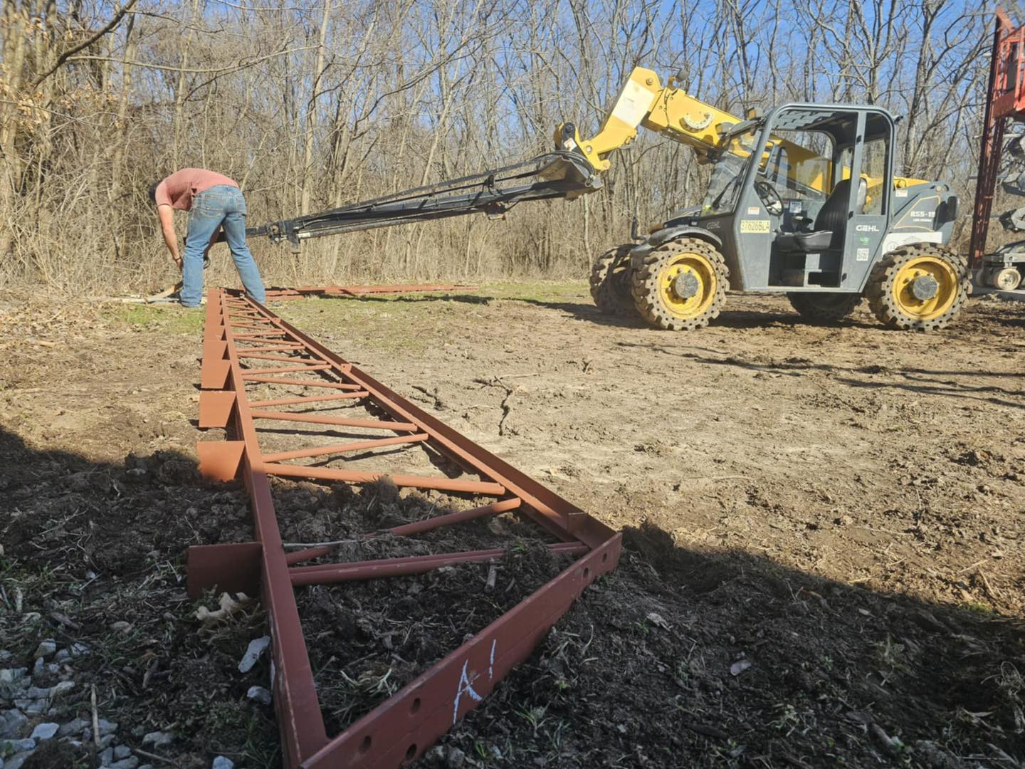Person placing red metal frame with telescopic handler's help, bare trees in Litchford background.