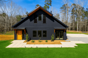 Modern black barndo with steep roof, large windows, wood doors, on lawn with trees and blue sky.
